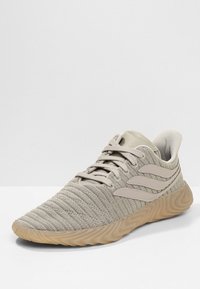 Ljusbeige stickad sneaker med vågig gummisula, tre sidostripes och snörning framtill på en helt vit bakgrund.