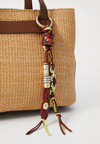 Tote en paille tissée avec des anses en cuir brun. Le sac présente un pompon décoratif tressé en marron, jaune et vert, agrémenté de perles et d'accents en métal.