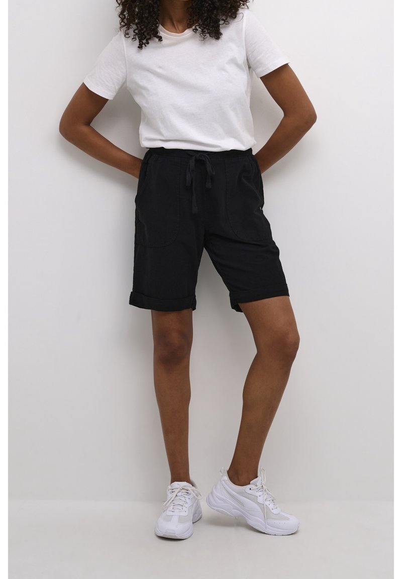 Pantalones cortos de algodón negros con puños enrollados, una cinturilla con cordón y dos grandes bolsillos frontales, combinados con una camiseta blanca de manga corta y zapatillas blancas.