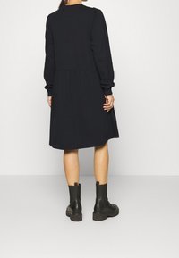 Svart långärmad klänning med en rynkad kjol; mjukt tyg; kombinerad med svarta ankelstövlar med en kraftig sula och dragflik.