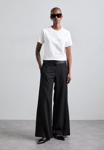 T-shirt en coton blanc avec un petit logo, associé à un pantalon large noir et des chaussures noires à bout pointu. Modèle portant des lunettes de soleil et des boucles d'oreilles.