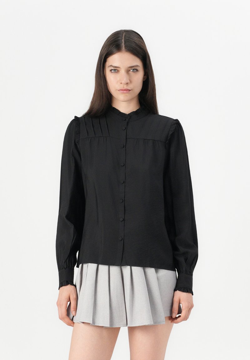 Camicia nera con bottoni, colletto arricciato e maniche lunghe, con dettagli plissettati sulle spalle e sui polsini. Tessuto dalla superficie liscia.