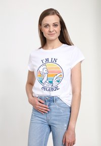 Hvid bomulds t-shirt med oprullede ærmer, der pryder et farverigt kyllingegrafik og teksten "JEG ER I KONTROL" mod en solnedgangsgradient baggrund.