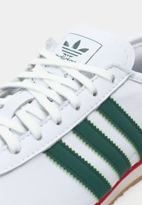 Zapatilla blanca de Adidas con rayas verdes, cordones blancos y logo de Adidas en la lengüeta, mostrando cuero texturizado y borde de suela roja.