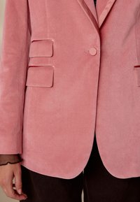 Pink velvet blazer with a single button closure, two front pockets, and a smooth texture. Features notched lapels and a tailored fit.