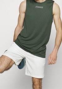 Zielona bezrękawnikowa koszulka sportowa z szarym logo, zestawiona z białymi szortami. Szorty mają boczne kieszenie i swobodny krój.