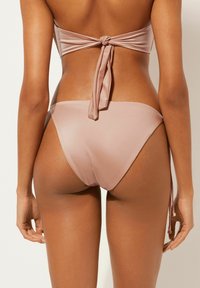 Jasnoróżowy bikini o gładkim satynowym wykończeniu. Górna część ma zawiązywane z tyłu zapięcie, a dolna część charakteryzuje się minimalistycznym wzornictwem i bocznymi trokami do regulacji.