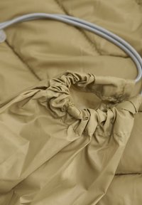 Beige, vadderad tyg med en samlad öppning. Har vit snodd för justering och en slät, glänsande yta med synliga sömmar.