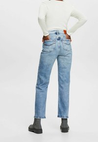 Jeans de mezclilla azul claro de pierna recta con dos bolsillos traseros y una etiqueta de cuero marrón en la cinturilla. Llevados con una camiseta blanca de canalé y botas negras.