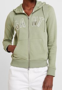 Sudadera con capucha verde con cremallera, hecha de tela suave, que presenta un bolsillo tipo canguro, capucha con cordón y letras grandes "GANT" en blanco.
