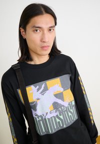 Schwarzes Langarmshirt mit einem großen grafischen Motiv in Lila, Gelb und Grau im Rodeo-Stil. Die seitlichen Ärmel haben ein Schachbrettmuster.