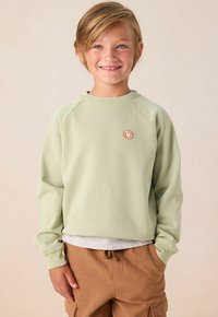 Sweatshirt vert clair avec des poignets côtelés, des manches raglan et un petit logo orange brodé sur la poitrine. Porté par-dessus une chemise grise et un pantalon marron.