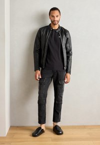 Giacca bomber in pelle nera su una t-shirt nera, abbinata a pantaloni cargo neri con tasche e scarpe slip-on nere. Sfondo in legno.