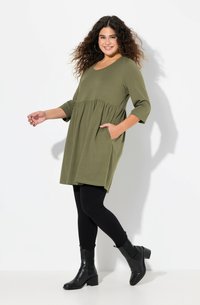Mujer sonriente con cabello rizado que lleva un vestido verde aceituna, leggings negros y botas negras hasta el tobillo, de pie con una mano en el bolsillo.
