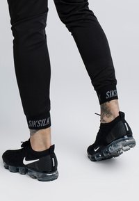 Schwarze Sporthosen mit Logo-Details an den Bündchen, kombiniert mit schwarzen Sneakers mit transparenten Sohlen und weißen Akzenten. Nahaufnahme der Unterschenkel.