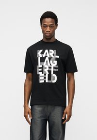 Νέος άνδρας που φοράει μαύρο T-shirt με λευκό παραμορφωμένο λογότυπο Karl Lagerfeld και σκούρο γκρι τζιν, στέκεται μπροστά σε απλό ανοιχτό φόντο.