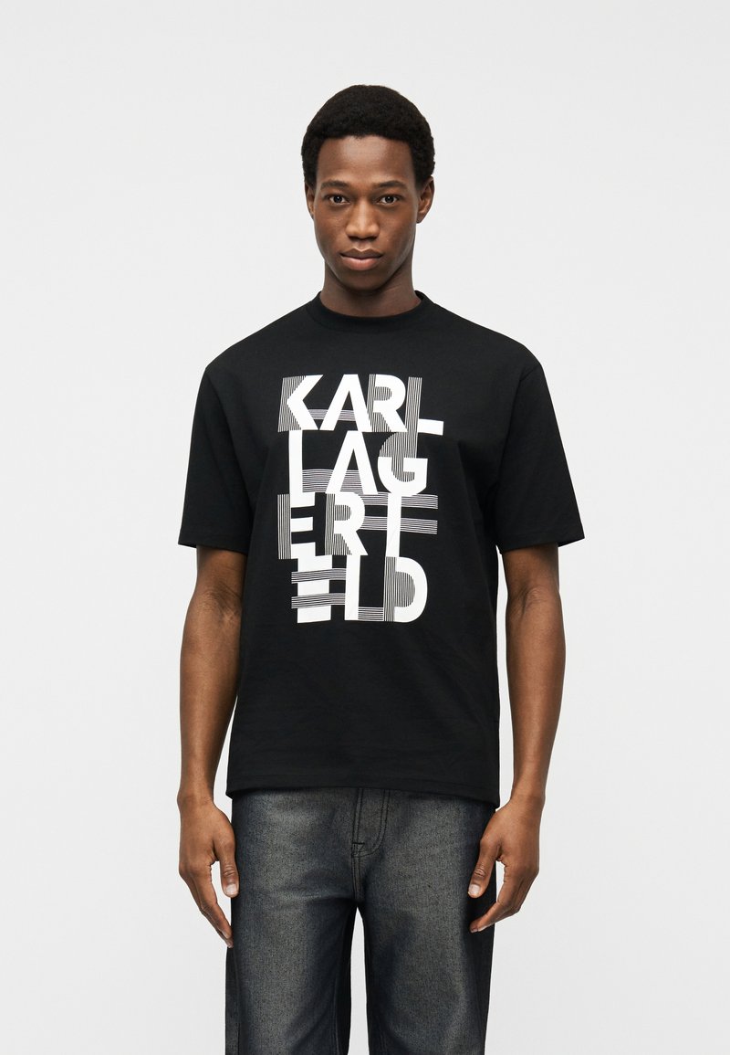 Νέος άνδρας που φοράει μαύρο T-shirt με λευκό παραμορφωμένο λογότυπο Karl Lagerfeld και σκούρο γκρι τζιν, στέκεται μπροστά σε απλό ανοιχτό φόντο.