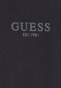 Navy blue fabric with ribbed texture featuring silver embroidered "GUESS" logo and "EST. 1981" below. Simple, clean design.