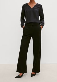 Blusa nera con scollo a V e maniche lunghe, abbinata a pantaloni neri a gamba larga. L'outfit presenta una texture liscia e un design elegante, completato da tacchi a punta.