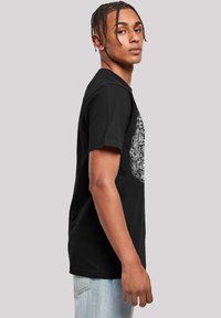 Black cotton t-shirt features short sleeves and a round neckline, with intricate white graphic design on the front. Denim jeans complete the look.
