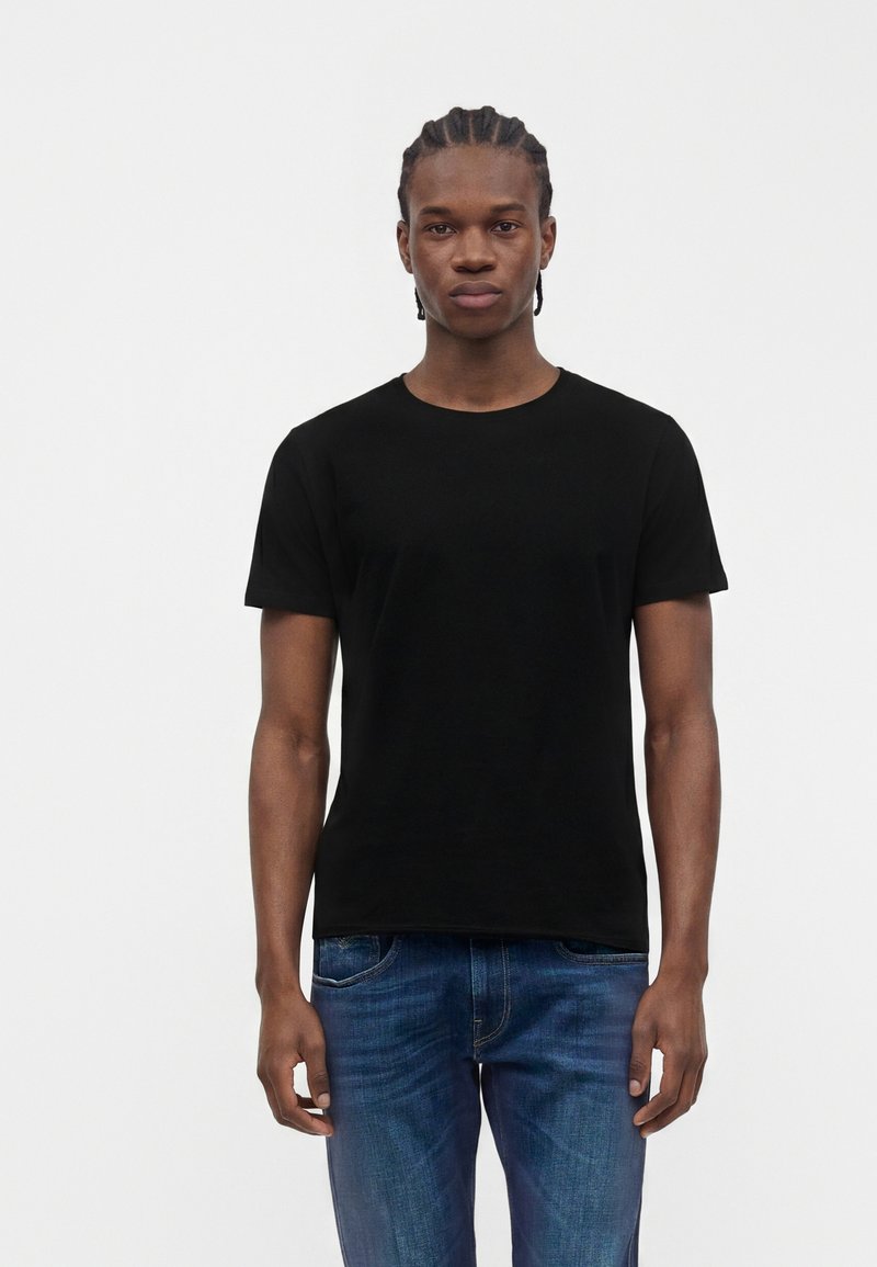 Jeune homme aux cheveux tressés, debout face à l’avant, portant un t-shirt noir uni et un jean bleu sur fond blanc.