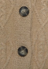 Beige Strickjacke mit einem strukturierten Zopfmuster und zwei großen, dunklen runden Knöpfen mit hellen Wirbeln und zwei Löchern.