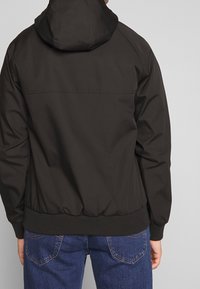 Chaqueta negra con capucha, de textura suave, puños elásticos y cintura ajustable. Presenta un diseño sencillo con una costura a lo largo de la espalda.