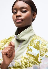 Γυναίκα με σκούρο δέρμα και κοντά μαλλιά κλείνει το φερμουάρ ενός ανοιχτοπράσινου φλις τζάκετ Columbia με σχέδια, κοιτάζοντας την κάμερα με αυτοπεποίθηση.