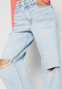 Jeans de mezclilla azul claro con un ajuste relajado, áreas de rodillas desgastadas y detalles de costura visibles. La cintura incluye herrajes de metal.