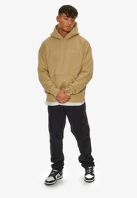 Beige hoodie with a kangaroo pocket and logo. Paired with black cargo pants featuring side pockets and white sneakers with black accents.