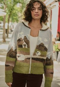 Knit cardigan in soft yarn with a light blue base, featuring a landscape design with houses and mountains in earthy tones. Zippered front, ribbed hem.