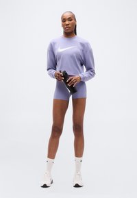 Μωβ φούτερ με λευκό λογότυπο Nike, σετ με ταιριαστό σορτς. Μαύρο παγούρι νερού στο δεξί χέρι, λευκές κάλτσες και αθλητικά παπούτσια.