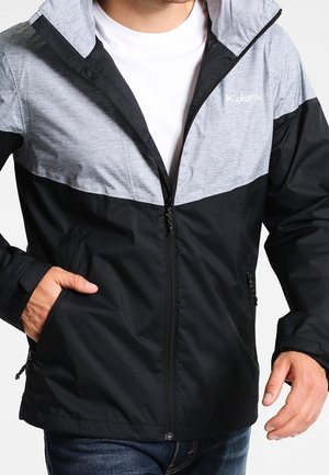 Man draagt een zwarte en lichtgrijze Columbia-jas met rits, met handen gedeeltelijk in de zakken, gecombineerd met een blauwe jeans en een wit shirt eronder.