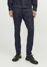 Jeans de denim azul oscuro con corte ajustado, que presentan costuras doradas en contraste y una cintura de altura media, acompañados de zapatos negros con cordones.