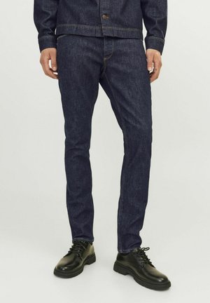 Dark blue denim jeans with slim fit, featuring contrasting gold stitching and a mid-rise waist, paired with black lace-up shoes.