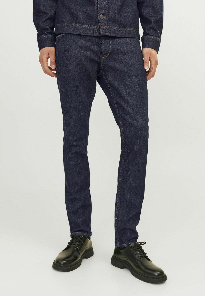 Jeans de denim azul oscuro con corte ajustado, que presentan costuras doradas en contraste y una cintura de altura media, acompañados de zapatos negros con cordones.