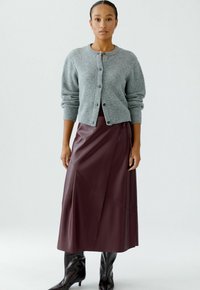 Grijze gebreide cardigan met knopen, gedragen over een bordeauxkleurige leren rok met hoekige naden, gecombineerd met hoogoplopende zwarte laarzen.