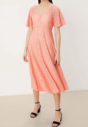 Kobieta w koralowo-różowej koronkowej sukience midi z krótkimi, falbaniastymi rękawami i dekoltem w kształcie litery V, w połączeniu z czarnymi sandałami na obcasie z paskiem wokół kostki.