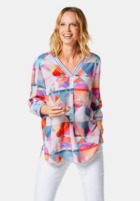 Blusa con motivo geometrico colorato, col v a V, maniche lunghe e polsini a righe. Il modello tiene un paio di occhiali; abbinata a pantaloni bianchi.