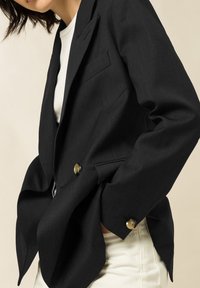 Blazer nero su misura con design monopetto, caratterizzato da due bottoni in ottone, tessuto leggermente strutturato e spalle strutturate, indossato sopra una camicia bianca.