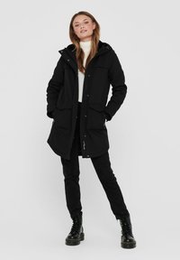 Parka noire avec capuche, fermeture éclair sur le devant, deux grandes poches avant et coupe décontractée. Portée avec un pantalon noir et des bottes de combat. Texture lisse.