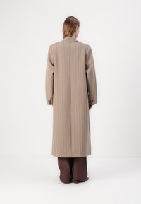Long brown coat with vertical light stripes, tailored fit, and wide sleeves. Worn over brown trousers, featuring button accents at the cuffs.