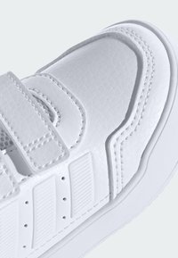 Baltas sintetinis sportbačius su tekstūruotu paviršiumi, lipniu dirželio užsegimu ir grioveliuotais akcentais. Turi siuvimo detales ir lygią pado dalį.