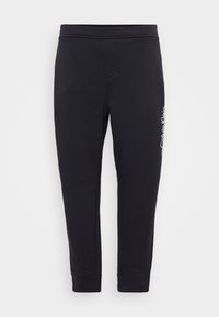 Schwarze Jogginghosen mit glatter Textur, elastischem Bund und Knöchellöchern. Mit einem weißen Logodetail auf der linken Seite.