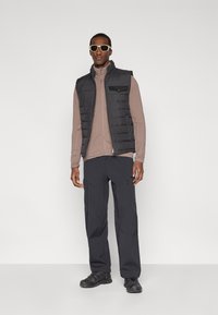 Zwarte doorgestikte bodywarmer over een lichtbruine lange mouwen shirt, gecombineerd met donkere broek en zwarte schoenen. Accessoires omvatten beige zonnebril.