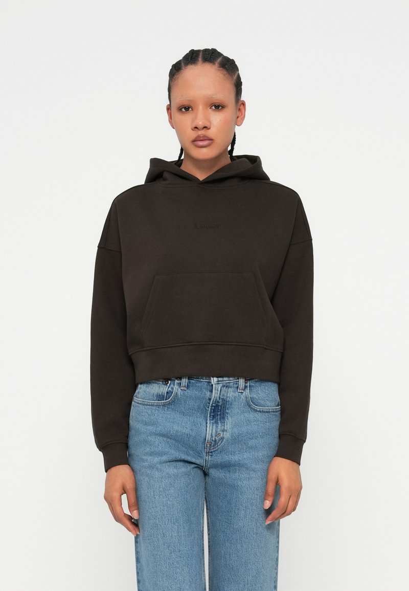 Sweatshirt cropped com capuz em castanho escuro, feito de tecido suave. Apresenta um bolso frontal e um logotipo discreto da marca no peito. Combinado com jeans azuis claros.
