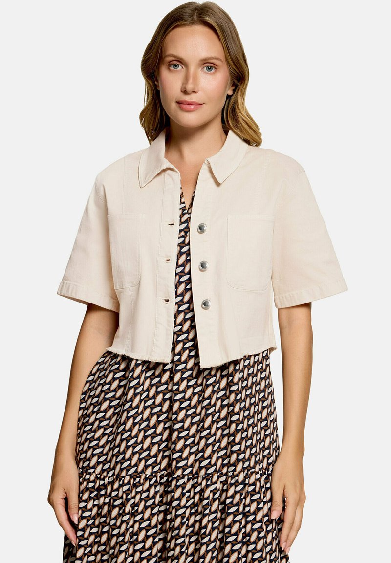 Femme aux cheveux mi-longs portant une veste courte beige à manches courtes par-dessus une robe à motifs avec des formes ovales.