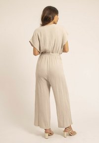 Combinaison tissée beige clair, à manches courtes, taille élastique, jambes larges, motif texturé, associée à des sandales à talons épais.