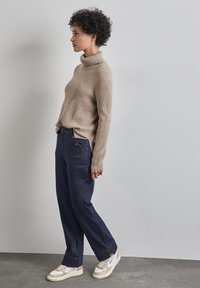 Un pull col roulé beige à côtes avec des manches longues, associé à un jean bleu foncé à jambes larges avec des boutons sur le côté et des baskets blanches.