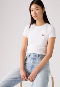 Biele tričko s krátkymi rukávmi a okrúhlym výstrihom, na hrudi s malým červeným logom Levi's, v kombinácii s svetlomodrými džínami so zvýšeným pásom.